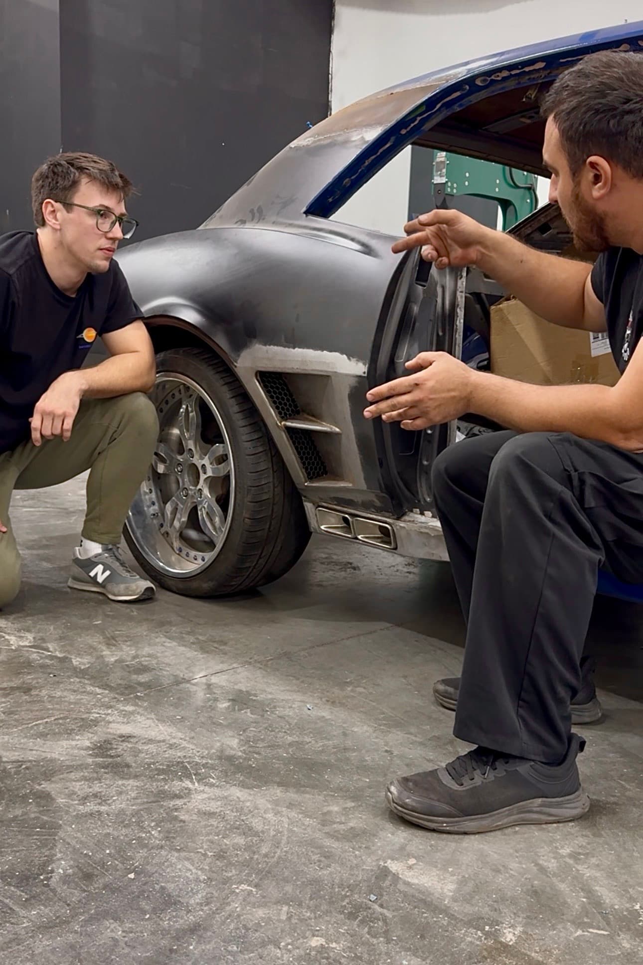 Edward Kolga in a shop, crouched at the rear wheel of a bare-metal build, going over the bodywork with the person building it