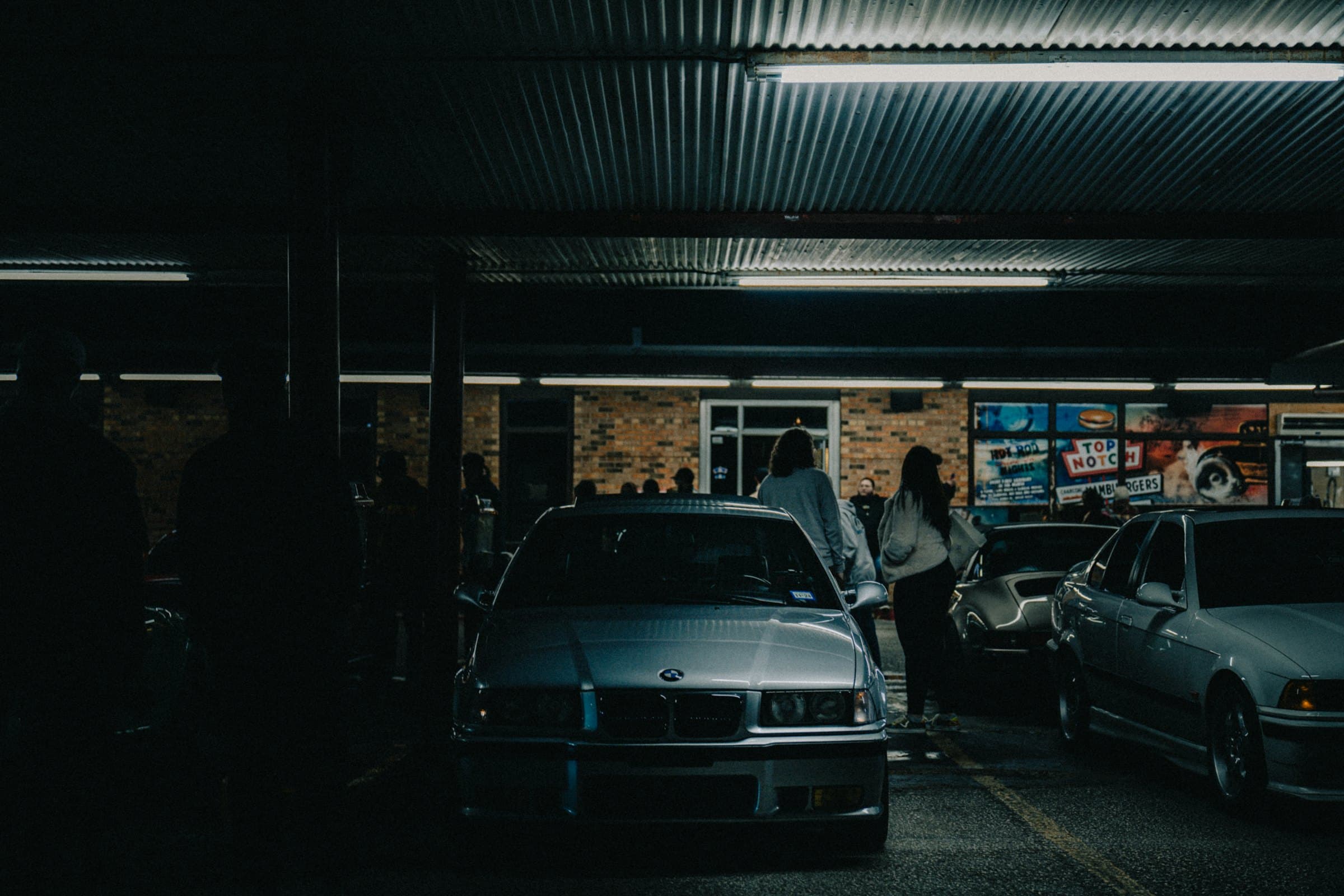 A night car meet under a lit canopy — an E36 BMW nose-on, people between the cars