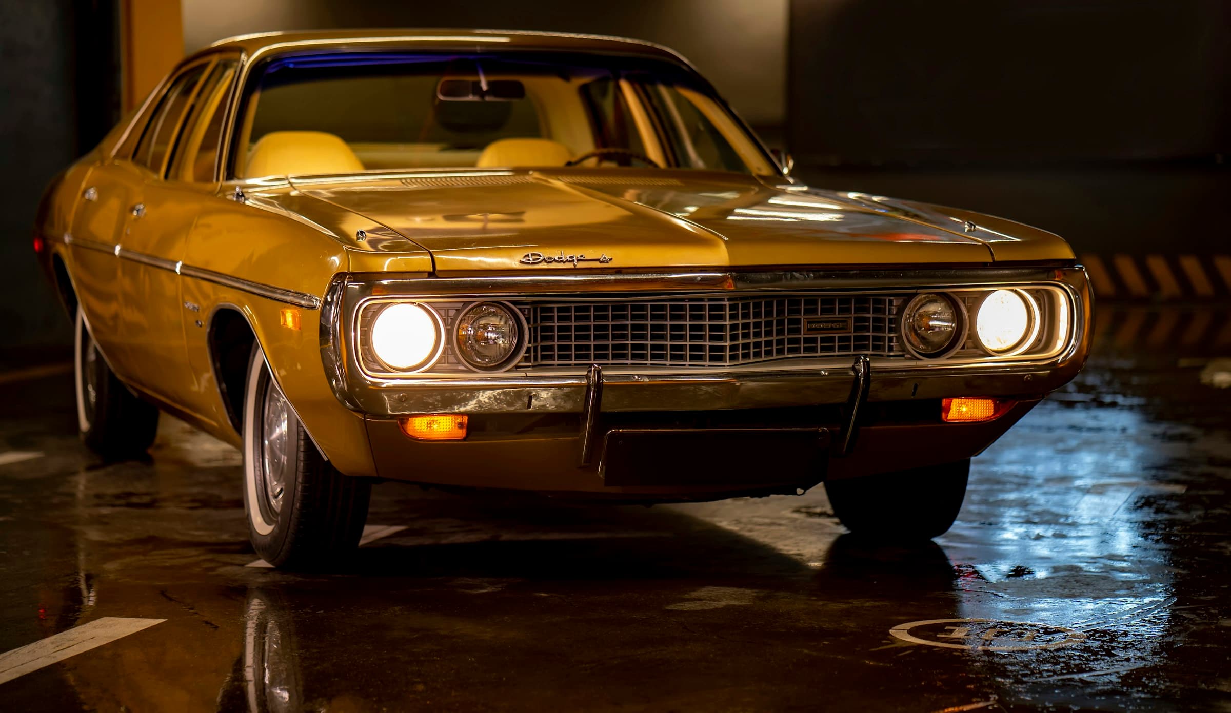 A gold 1970s Dodge sedan, headlights on, in a wet underground garage